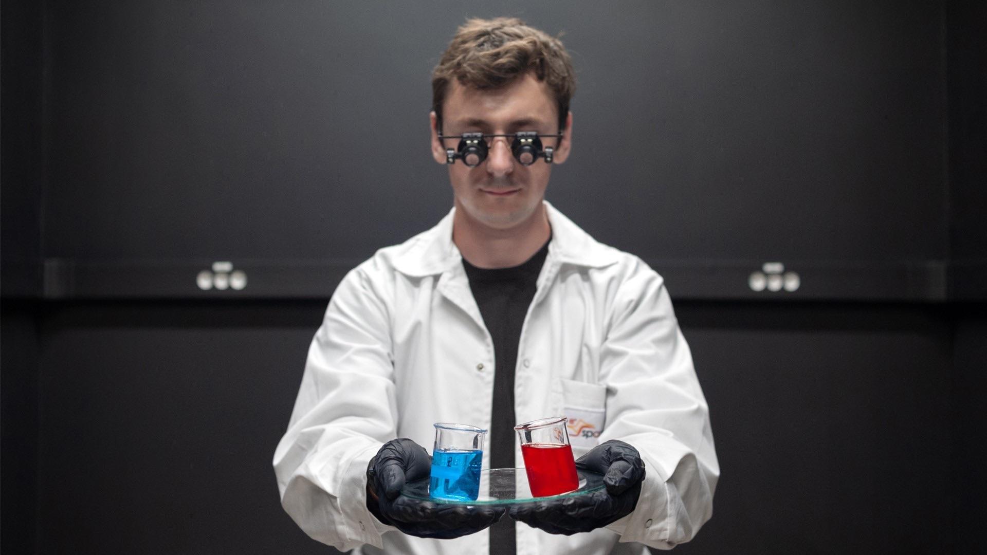 Grafika przedstawiająca naukowca w kitlu trzymającego kolby z niebieską i czerwoną cieczą